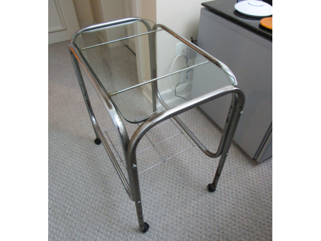 Mesa lateral cromada com tampo de vidro, estrutura arredondada e pés com rodinhas. 60 x 55 x 35 cm. - Anos 80